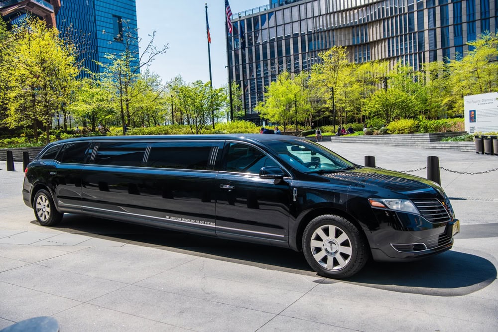 A long black stretch limousine from FL Limo Service Jacksonville is parked on a city street with modern buildings and green trees in the background on a sunny day.