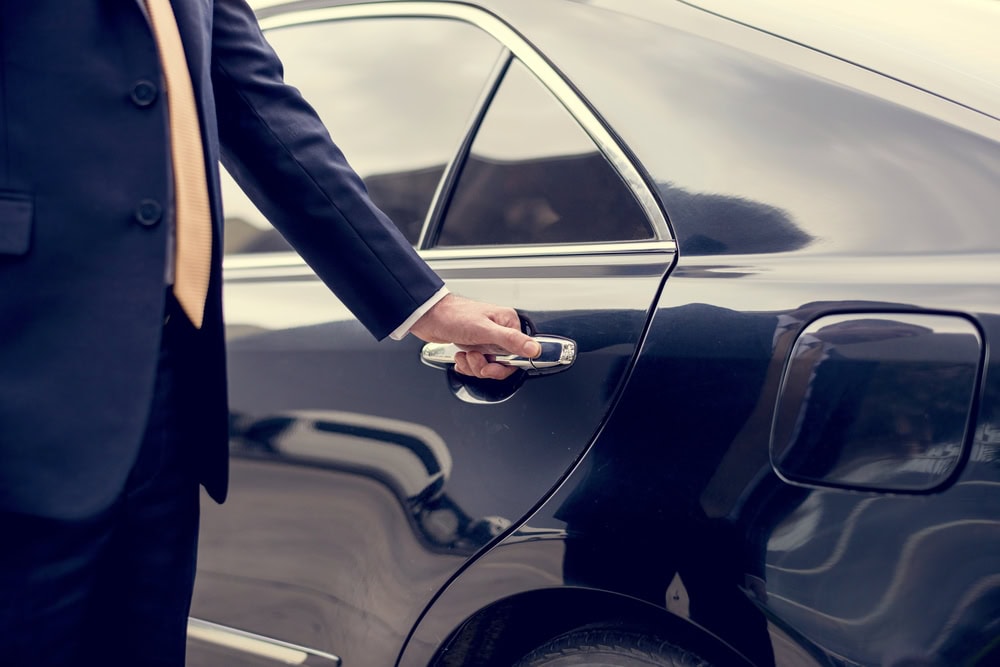 A person in a suit and tie is opening the rear door of a black car, with their hand on the door handle, suggesting a professional Limo Service Jacksonville, FL experience.