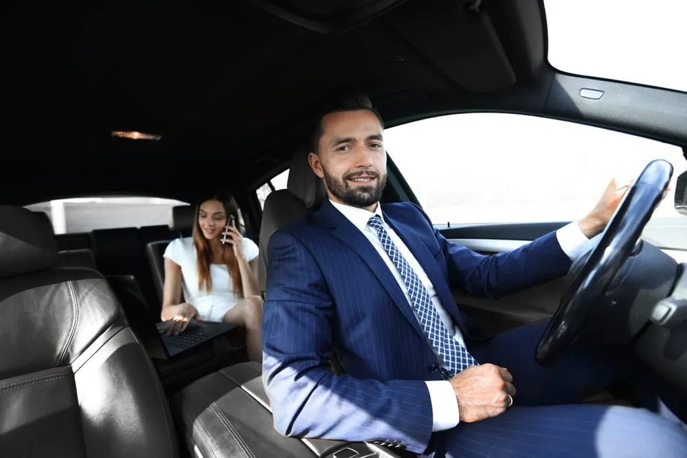 A man in a suit is driving a car for a limo service Jacksonville, smiling at the camera. In the back seat, a woman in business attire talks on the phone and uses her laptop, focused on work during her FL commute.
