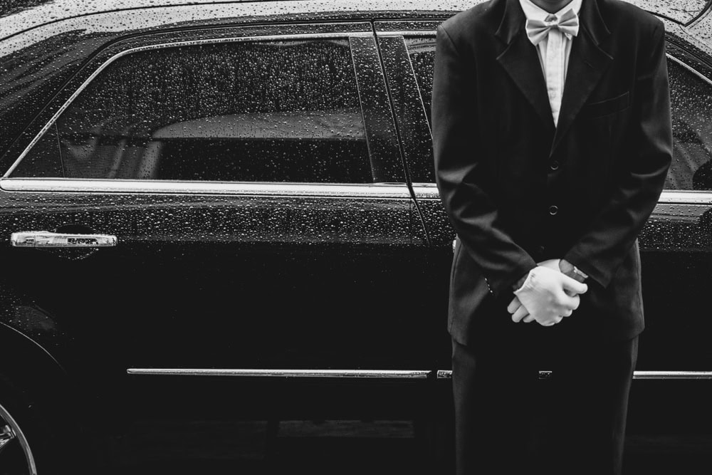 A person in a suit and bow tie stands with hands clasped in front of a black Limo Service Jacksonville car on a rainy FL day. Water droplets cover the car’s windows and door, while the person’s head is partially out of frame.