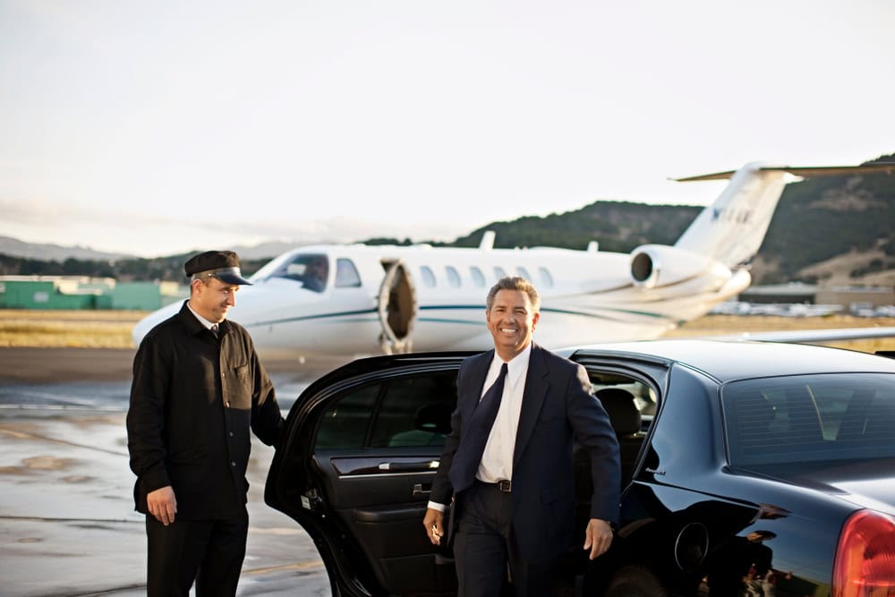A smiling man in a suit exits a black car while a chauffeur from a limo service Jacksonville, FL, holds the door open. A private jet is parked in the background on an airport runway.