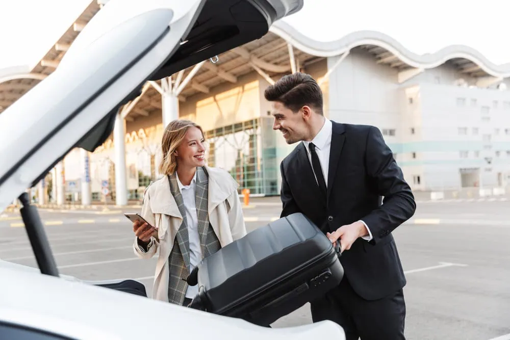 A smiling woman holding a phone stands next to a man in a suit, who is placing a suitcase in the trunk of a white car from a limo service Jacksonville, outside a modern FL airport terminal.