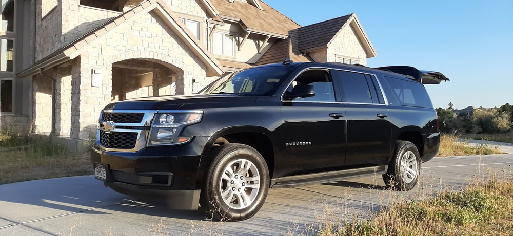 A black Chevrolet Suburban SUV, part of a Limo Service Jacksonville, FL fleet, is parked on a driveway in front of a large stone house. The rear hatch is open and the sun casts shadows on the driveway and grass nearby.