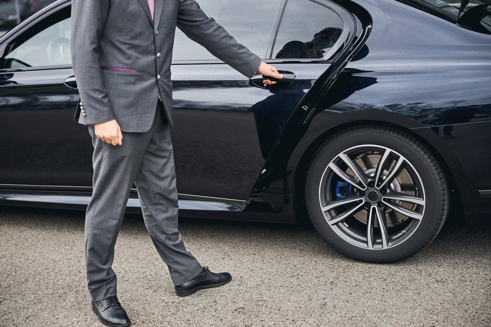 A person wearing a gray suit and black shoes is opening the rear door of a black luxury car from a Limo Service Jacksonville, FL, parked on a paved surface. The person's face is not visible.