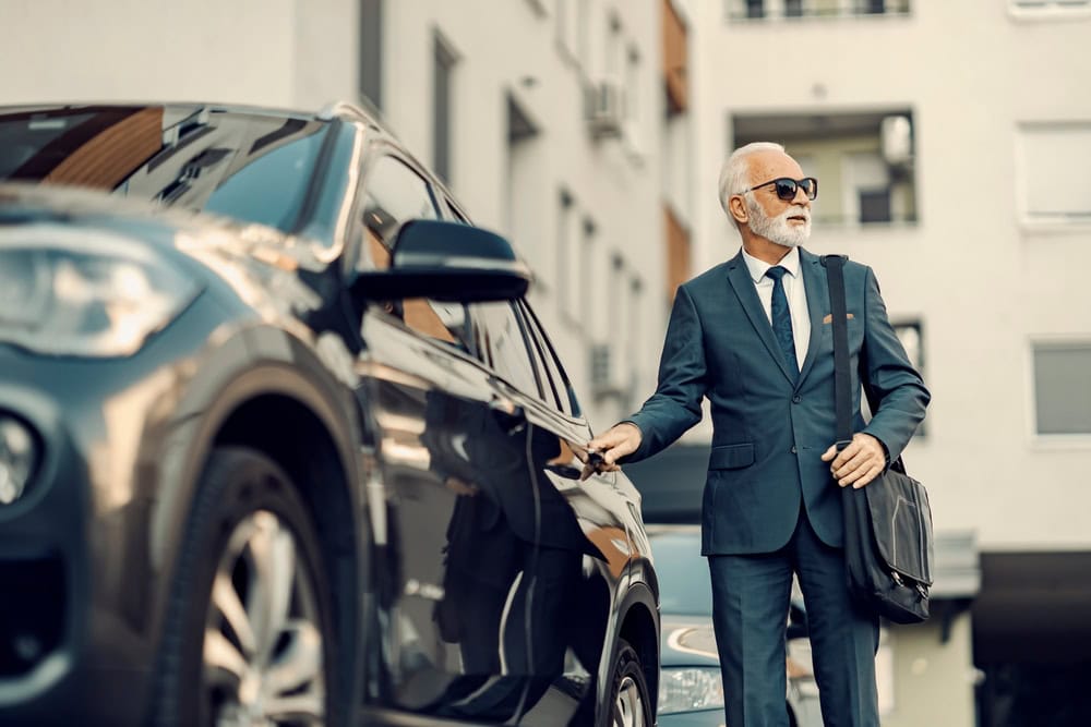 An older man in a suit and sunglasses stands next to a parked car, holding a briefcase and opening the door—a classic scene of professionalism with Limo Service Jacksonville, FL on a bustling city street.