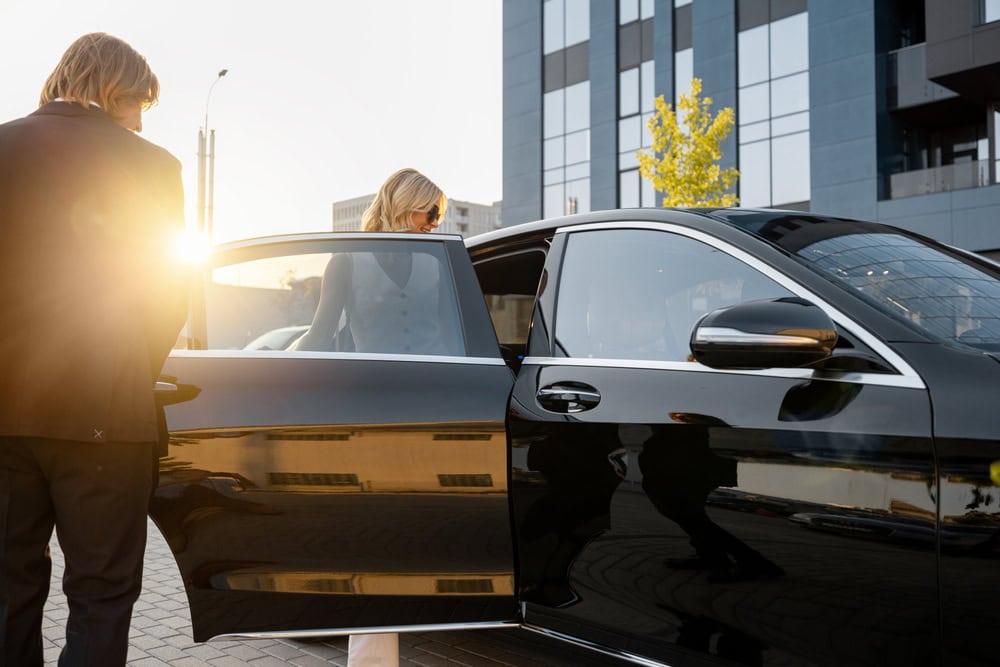 A person is getting into the back seat of a black car provided by Limo Service Jacksonville, while another stands nearby. The sun is shining brightly, with a modern building in the background, capturing a classic day in FL.