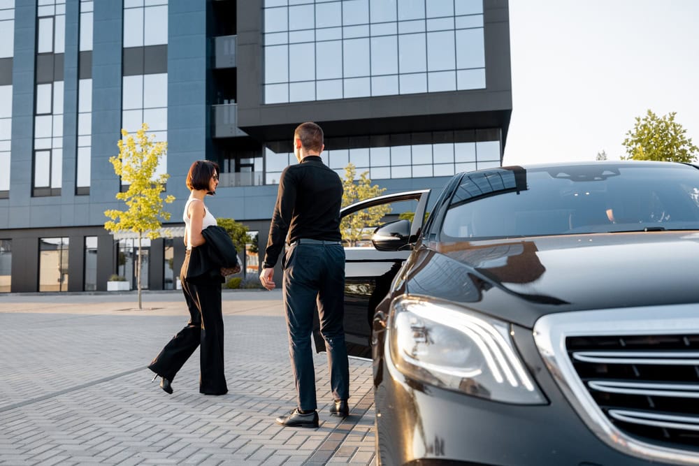 A man opens a car door for a woman outside a modern office building on a sunny day. Both, dressed in formal attire, stand near the sleek vehicle provided by Limo Service Jacksonville, FL.