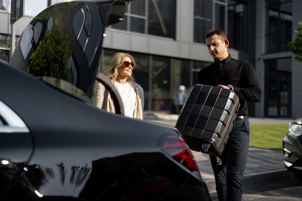 A man loads a black suitcase into the trunk of a parked car from a FL Limo Service Jacksonville while a smiling woman wearing sunglasses stands nearby outside a modern glass building.