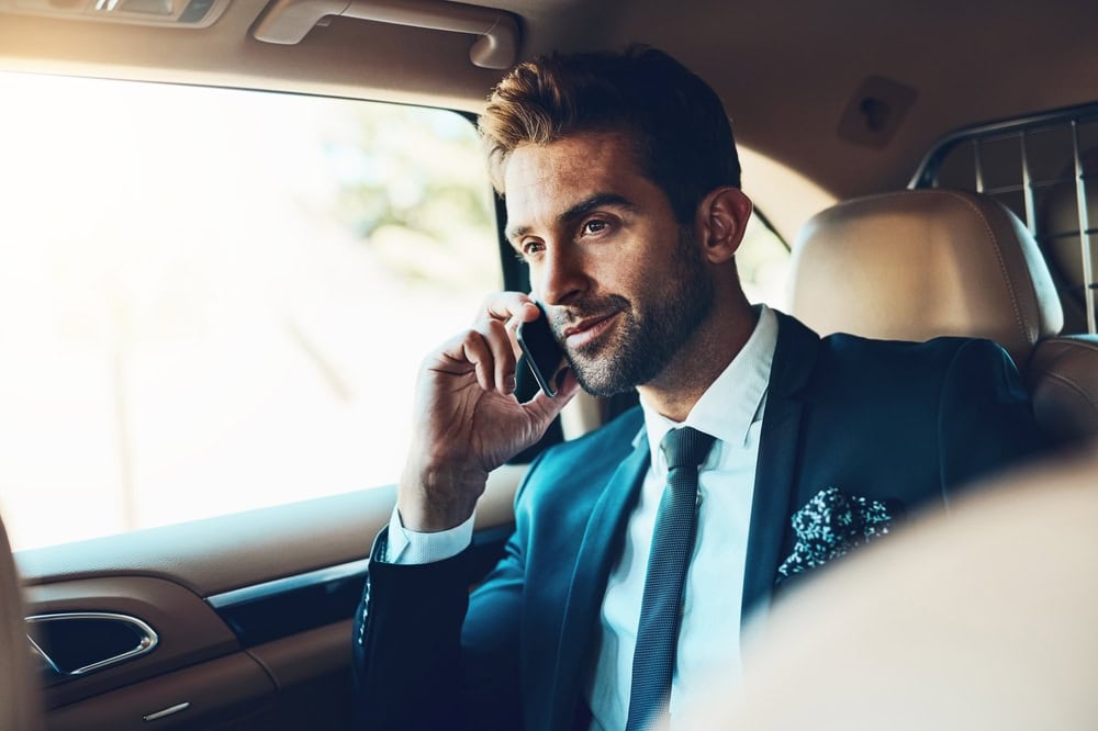 A man in a suit sits in the backseat of a luxury car from a Limo Service Jacksonville, smiling while talking on his mobile phone. Sunlight streams through the window, highlighting his confident expression as he travels through FL.
