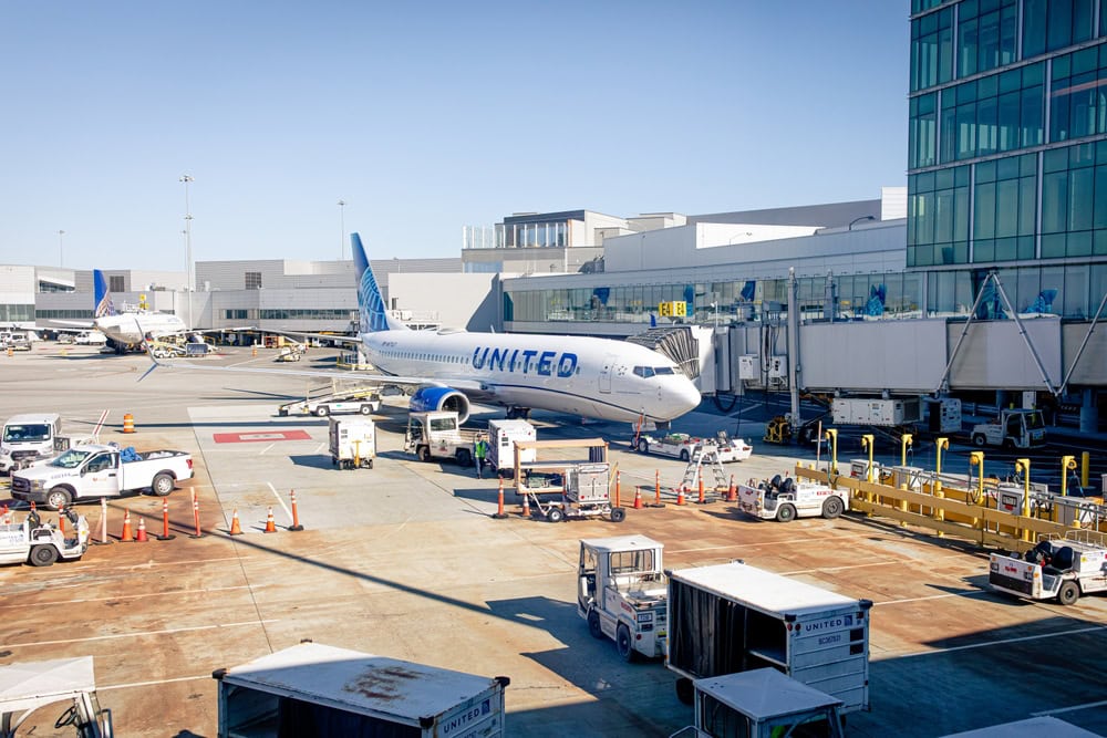 A United Airlines plane is parked at an airport gate in FL, surrounded by service vehicles and equipment. The terminal building and clear blue sky are visible in the background, with a Limo Service Jacksonville nearby for arriving passengers.