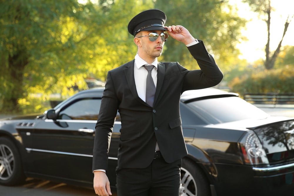 A man in a black suit, tie, and chauffeur cap stands in front of a black luxury car from a premium Limo Service Jacksonville, adjusting his sunglasses. Trees and sunlight are visible in the background of FL.