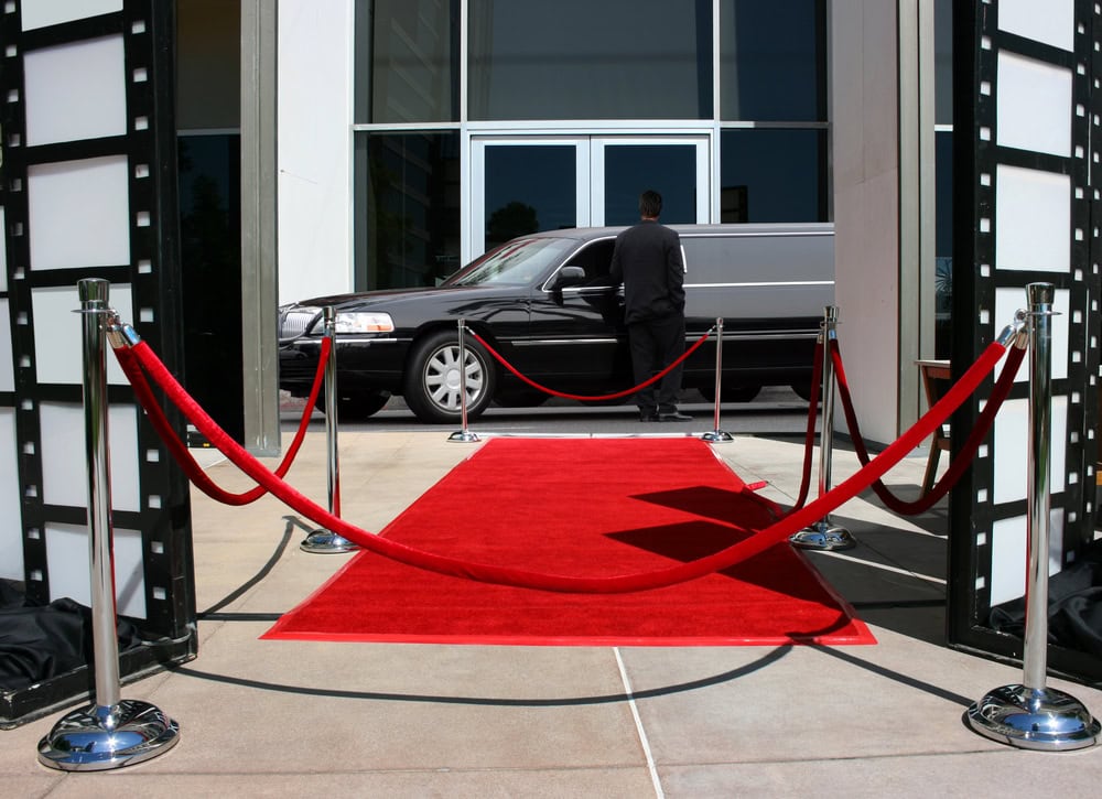 A red carpet stretches between velvet ropes toward a building entrance, with a black limousine from Limo Service Jacksonville parked in front and a person in formal attire standing nearby, capturing the luxury of VIP arrivals in FL.