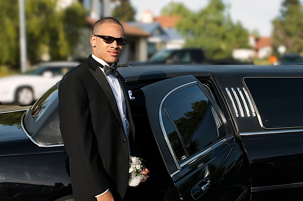 A young man in a tuxedo and sunglasses stands beside an open limousine door, holding a small bouquet—classic style meets FL elegance with Limo Service Jacksonville. Houses and greenery blur softly in the background.
