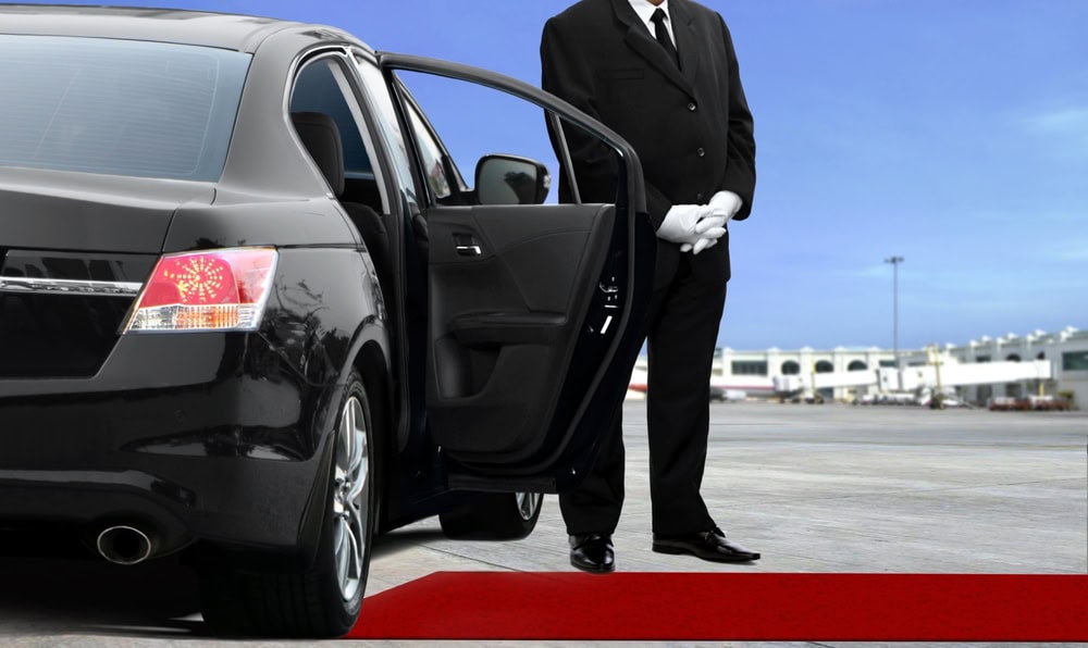 A chauffeur in a black suit and white gloves stands by an open car door from a Limo Service Jacksonville vehicle, next to a red carpet on an airport tarmac in FL, with the terminal building visible in the background.