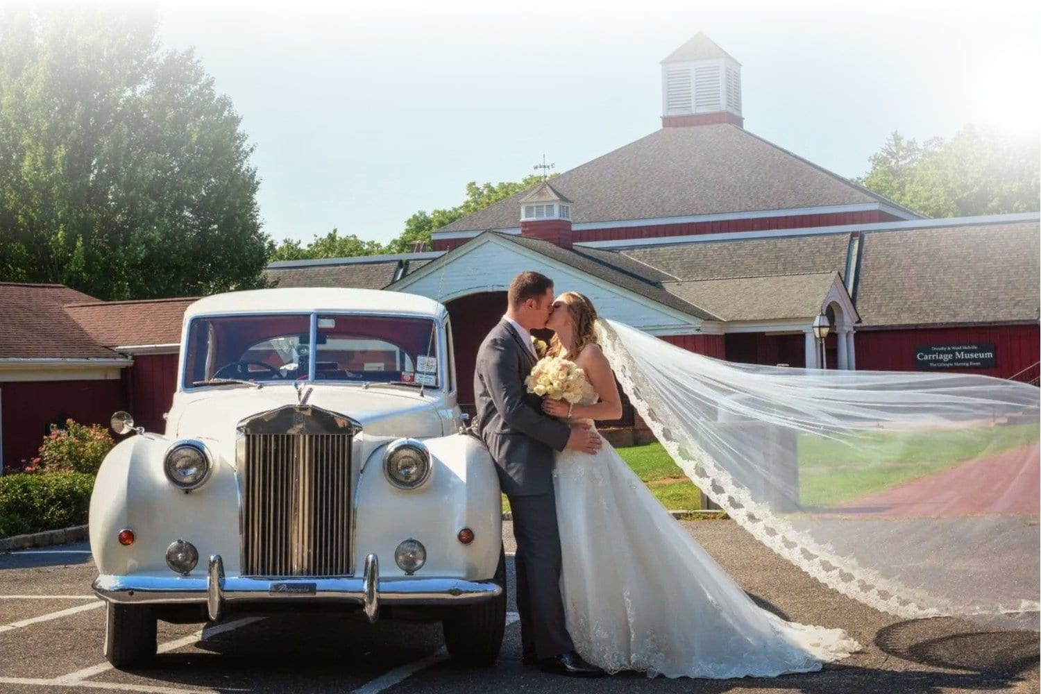 A bride and groom kiss beside a classic white car, with the bride’s long veil flowing in the wind. They are in front of a red barn-style building on a sunny day.