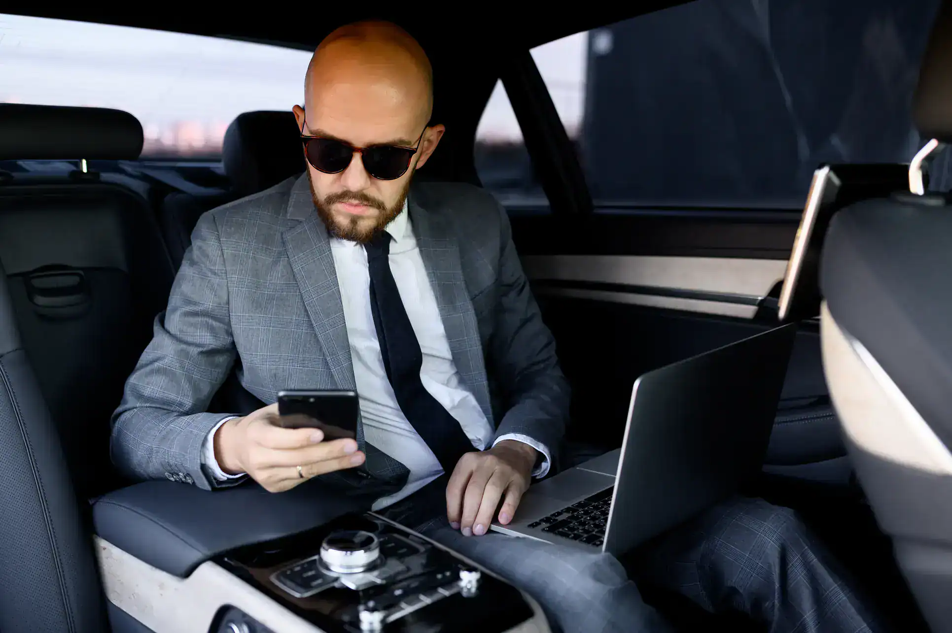 A man in a gray suit and sunglasses sits in the back seat of a car, looking at his smartphone. He has a laptop on his lap and appears focused, suggesting he is working while traveling.