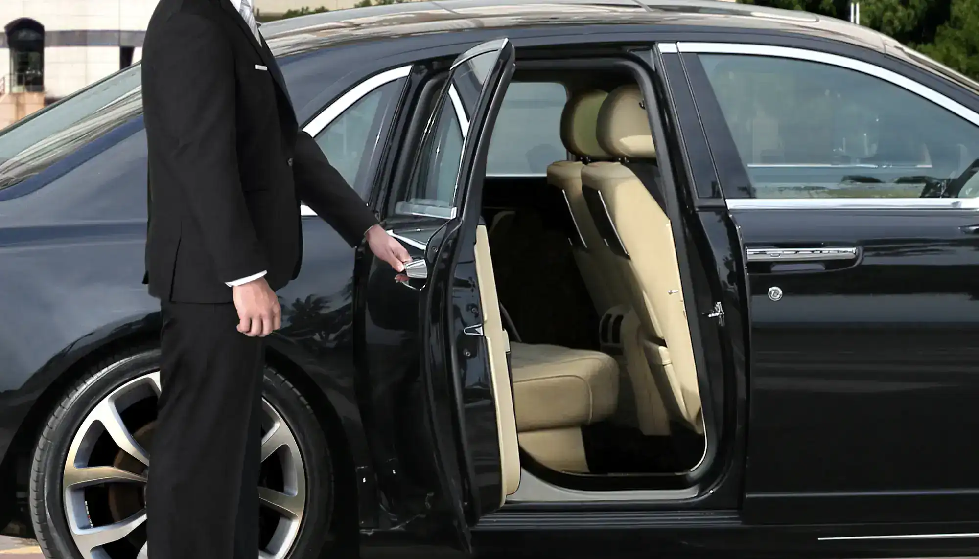 A person in a black suit stands next to a luxury car with the rear passenger door open, revealing beige leather seats inside.