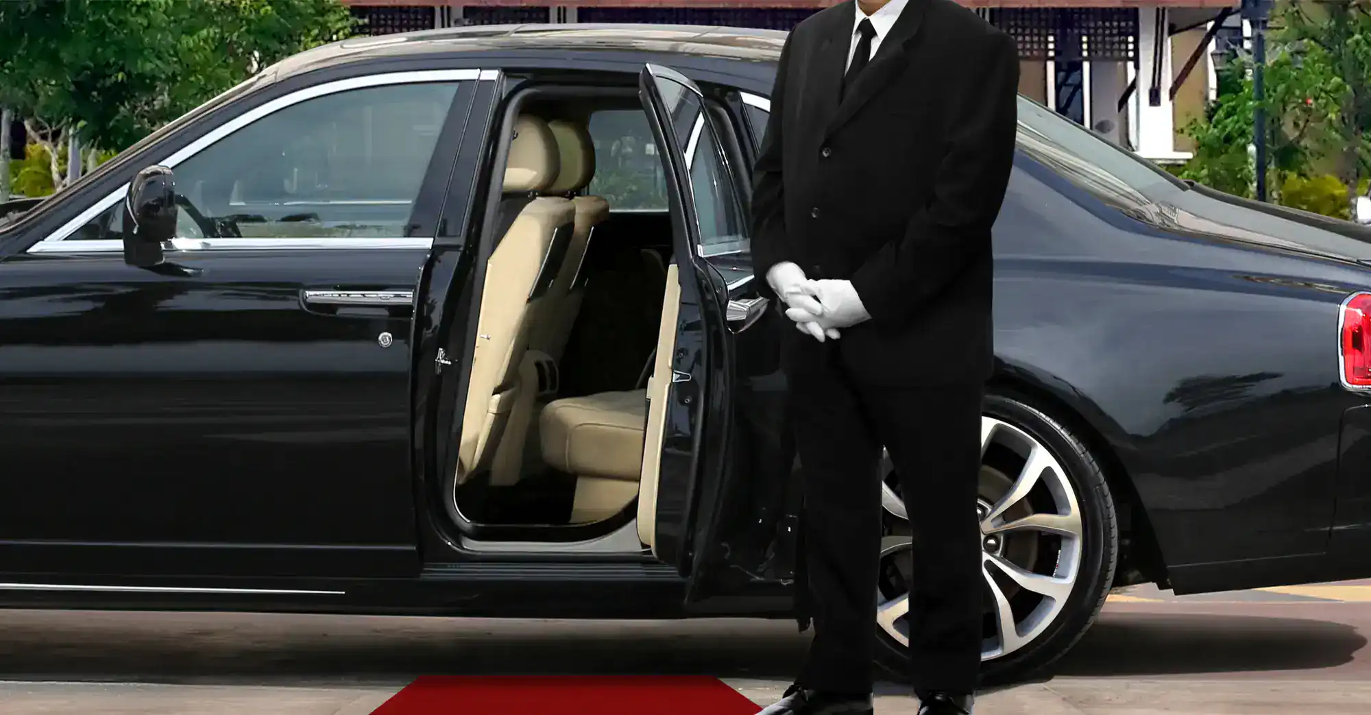 A chauffeur in a black suit and white gloves stands beside a luxury black car with an open back door, ready to assist passengers onto a red carpet.