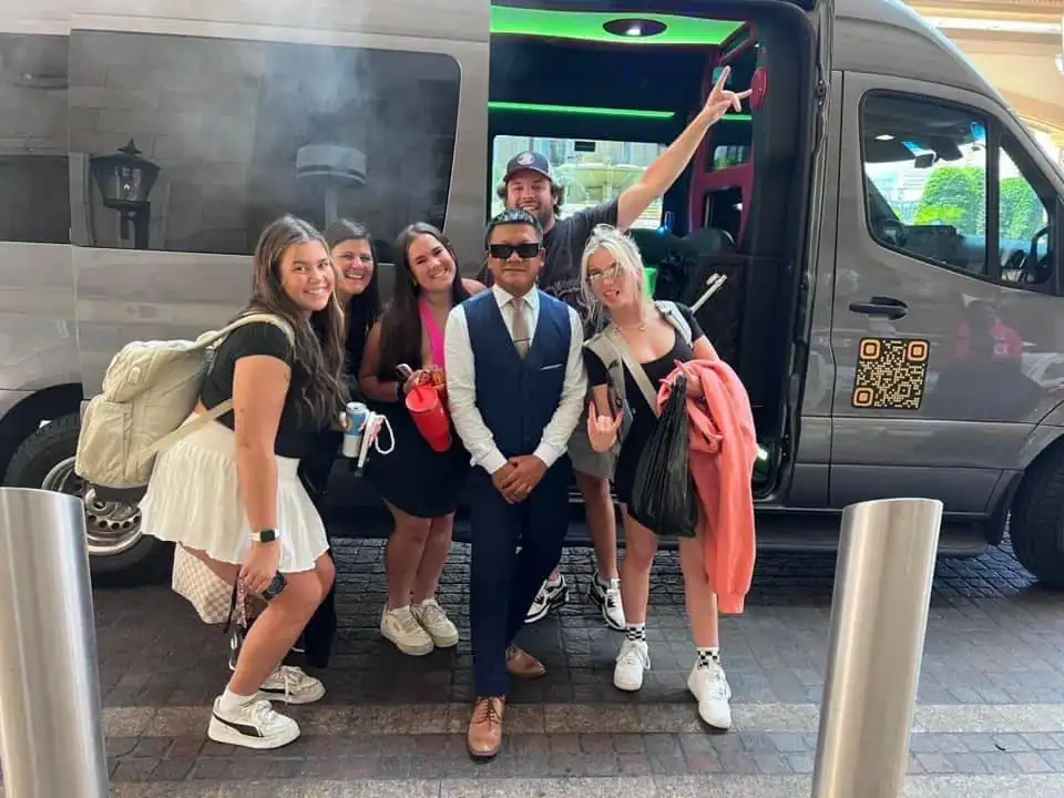 A group of five smiling people, four women and one man in casual clothes, pose with a suited man in front of a black van with an open door. Some hold bags and jackets; one person flashes a peace sign.