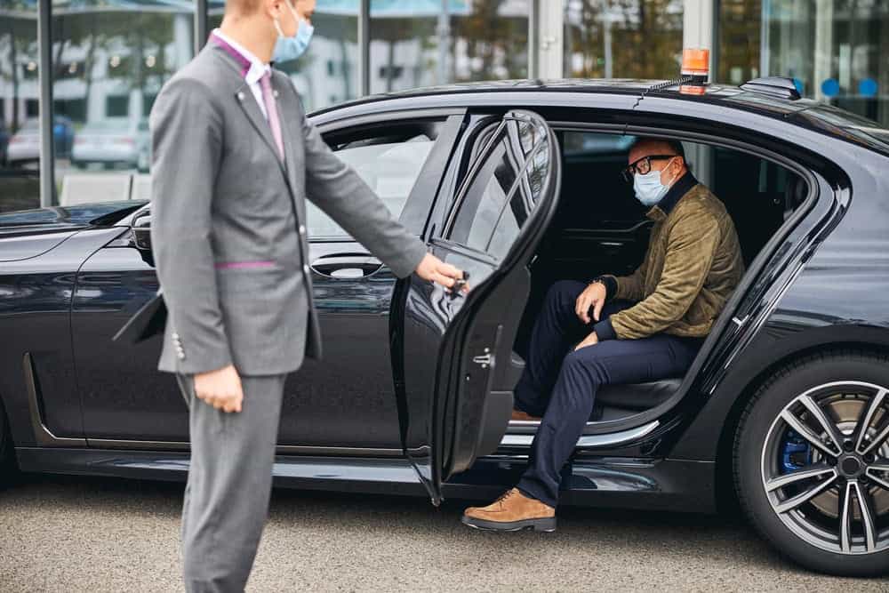 A man wearing a mask sits in the back seat of a black car, while another masked man in a suit holds the car door open for him. Both are outside, near a modern building.