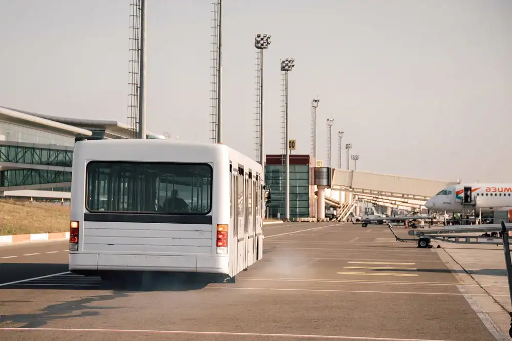 A white airport shuttle bus drives on the tarmac toward a modern terminal, with airplanes parked nearby and tall light poles lining the area, offering convenient transport similar to a Limo Service New Jersey on a clear day.