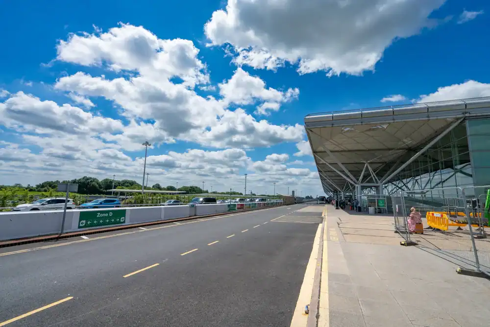 A wide view of an airport terminal exterior on a sunny day, featuring a glass building on the right, cars&mdash;including a Limo Service New Jersey vehicle&mdash;along the curb, blue sky, and scattered clouds overhead.