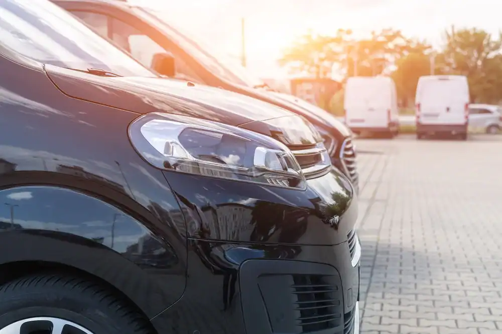 Row of parked black vans in a parking lot, sunlight shining in the background and white vans visible in the distance. The photo highlights the front headlights and grilles&mdash;perfect for showcasing a premium Limo Service New Jersey fleet.