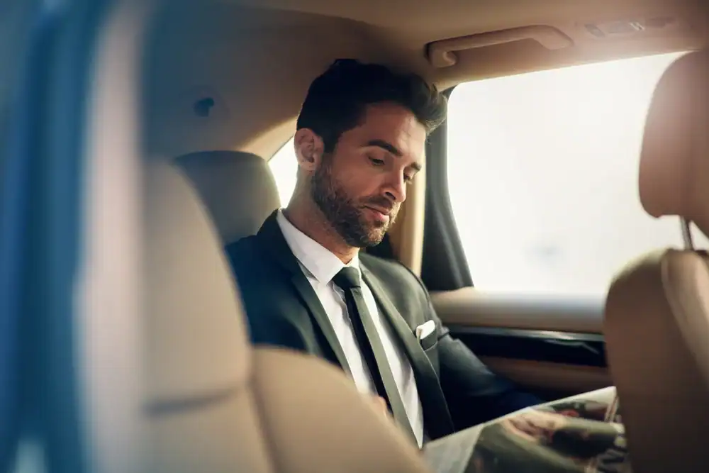 A man in a suit sits in the backseat of a car, likely enjoying a premium Limo Service New Jersey, gazing down at something out of view as sunlight streams through the window.