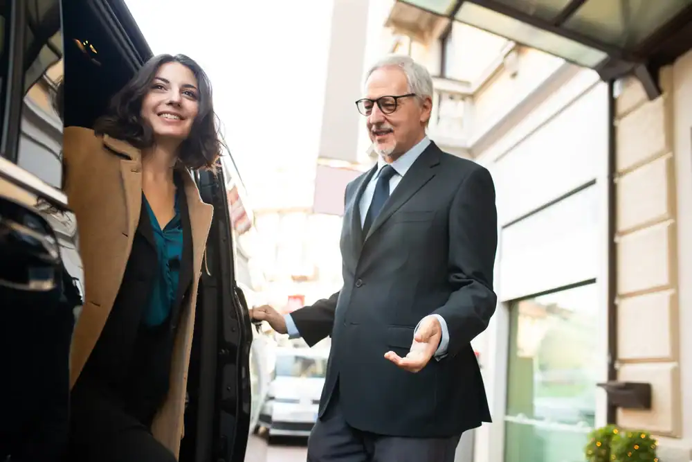 A smiling woman steps out of a car while a man in a suit and glasses, representing Limo Service New Jersey, stands nearby, holding the door open for her on a city street.