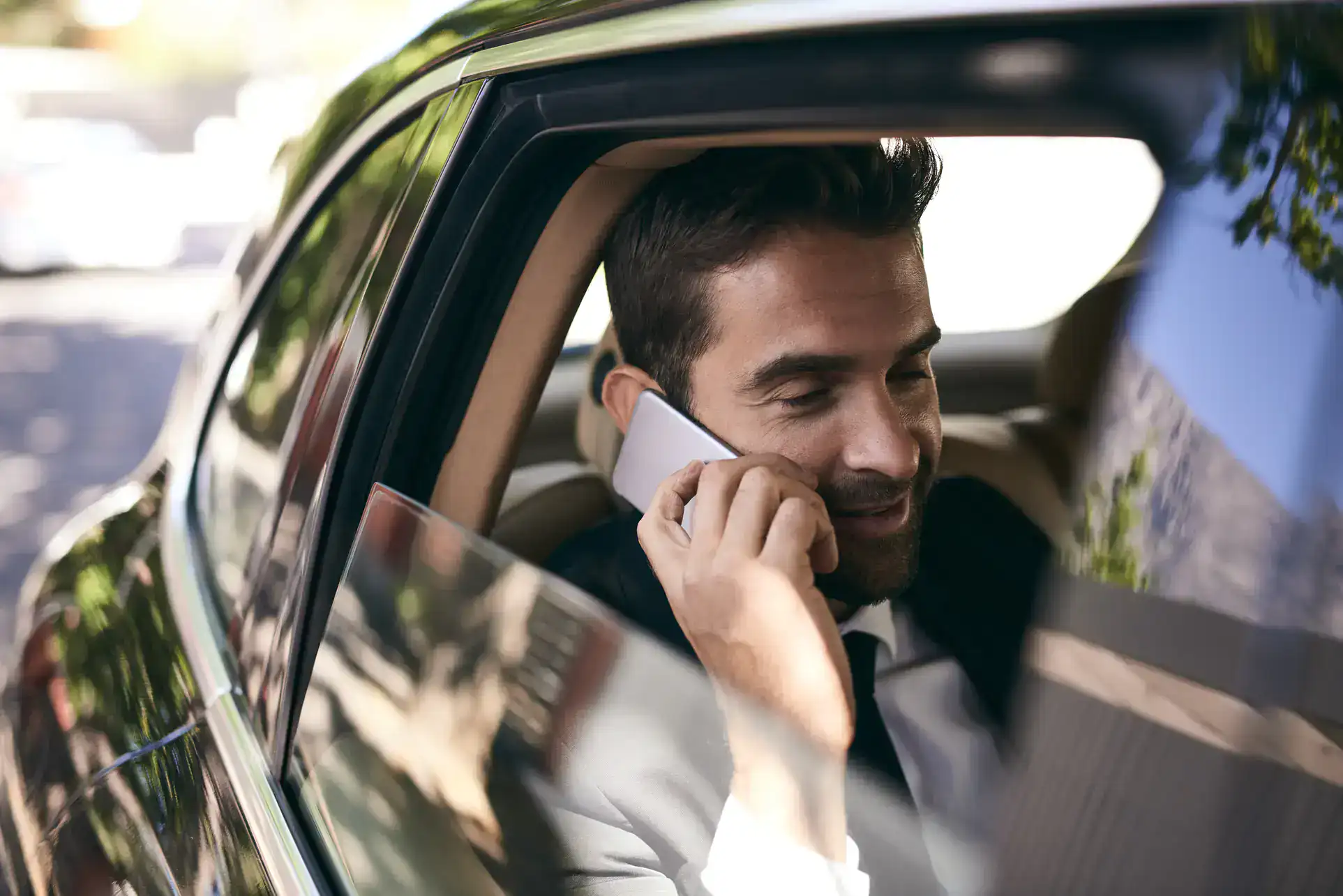 A man in a suit sits in the back seat of a Limo Service New Jersey car, smiling while talking on his smartphone. Sunlight reflects off the car window, and trees are visible outside.