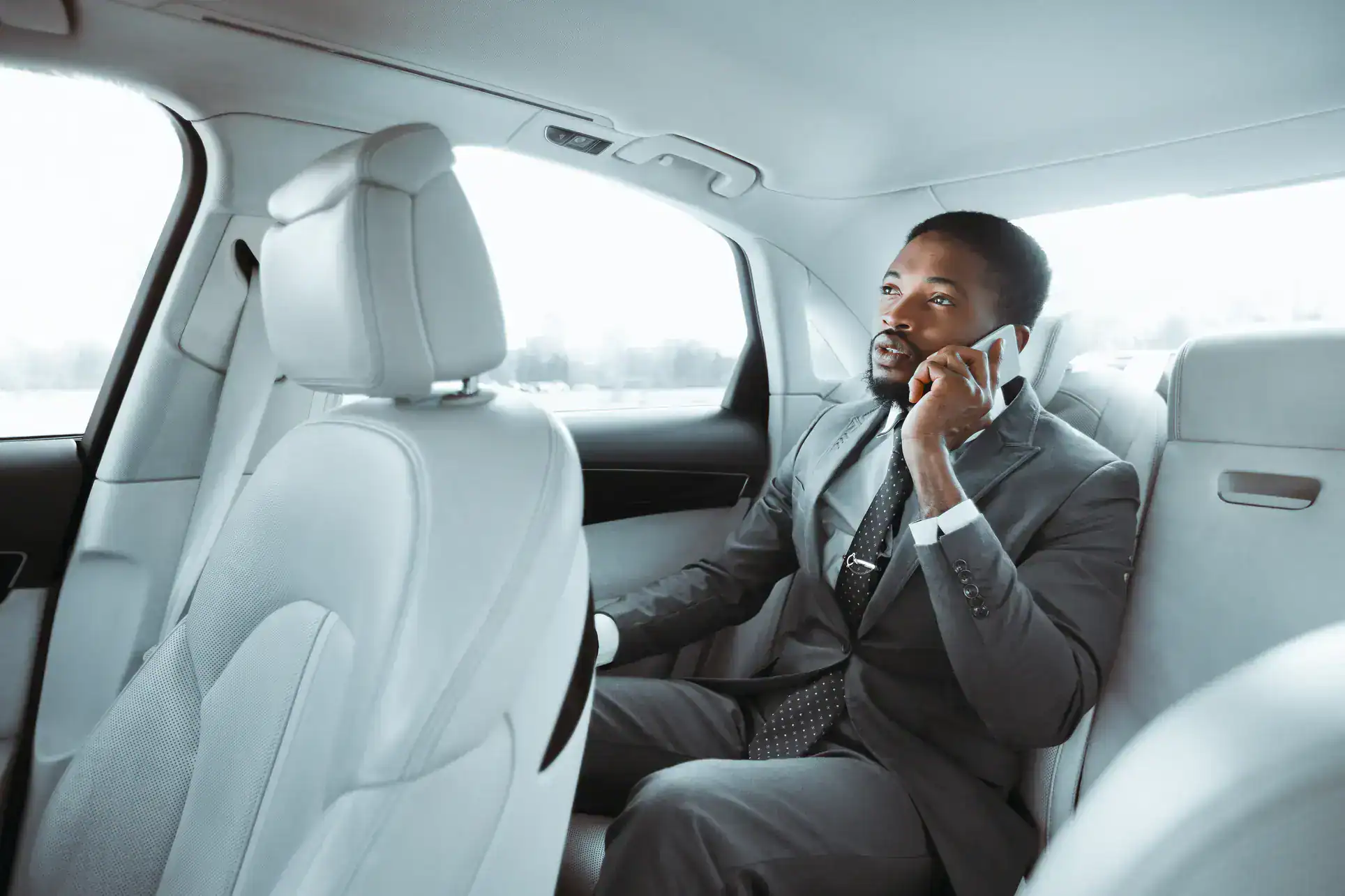 A man in a suit sits in the back seat of a car, talking on his cellphone. He looks out the window, appearing focused and engaged in conversation, enjoying the comfort of a modern, light-colored Limo Service New Jersey interior.