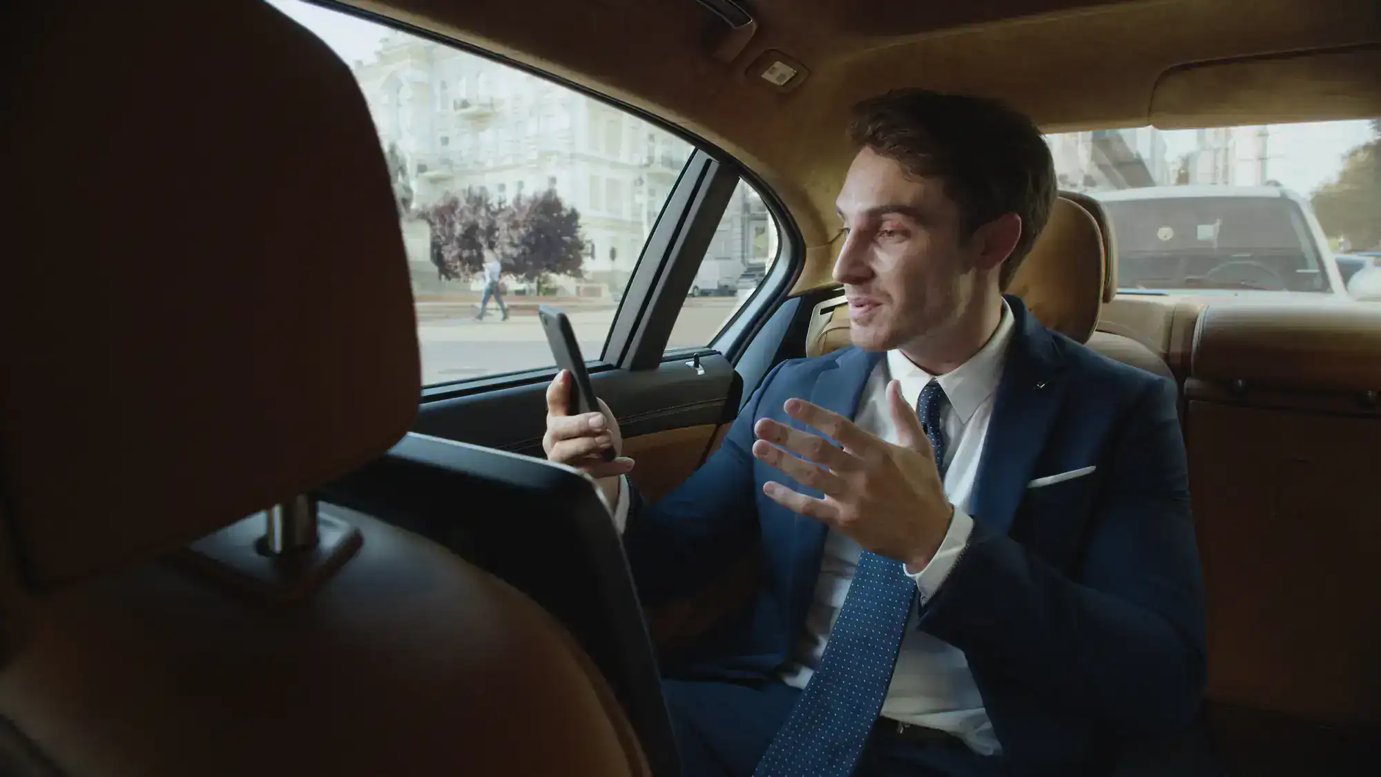 A man in a suit sits in the back seat of a Limo Service New Jersey car, holding a smartphone and gesturing as if on a video call, with city buildings visible through the window.