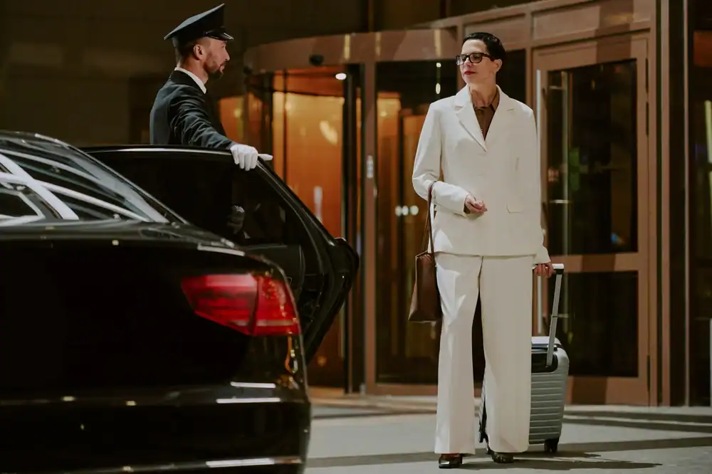 A chauffeur from a Limo Service New Jersey opens a car door for a woman in a white suit with glasses, holding a brown bag and pulling a suitcase outside a building with glass doors at night.