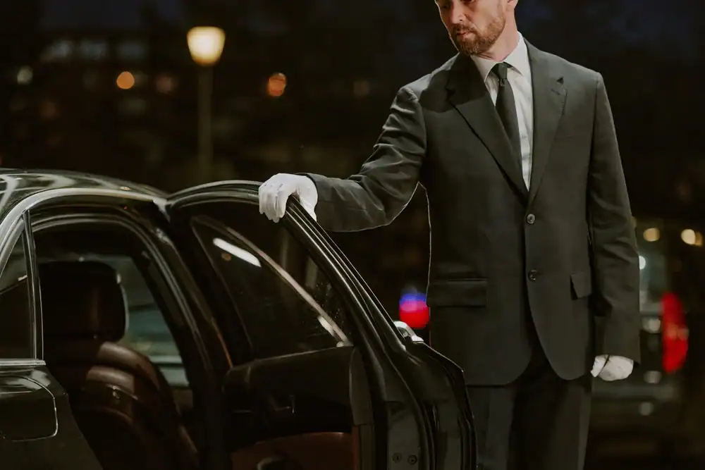 A man in a suit and white gloves stands next to a black car at night, holding the rear door open for a passenger&mdash;exemplifying premium Limo Service New Jersey.