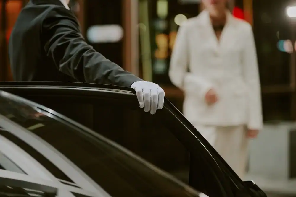 A person in a suit and white gloves holds open a car door, showcasing the professionalism of Limo Service New Jersey, while another person in a white outfit approaches in the blurred background.