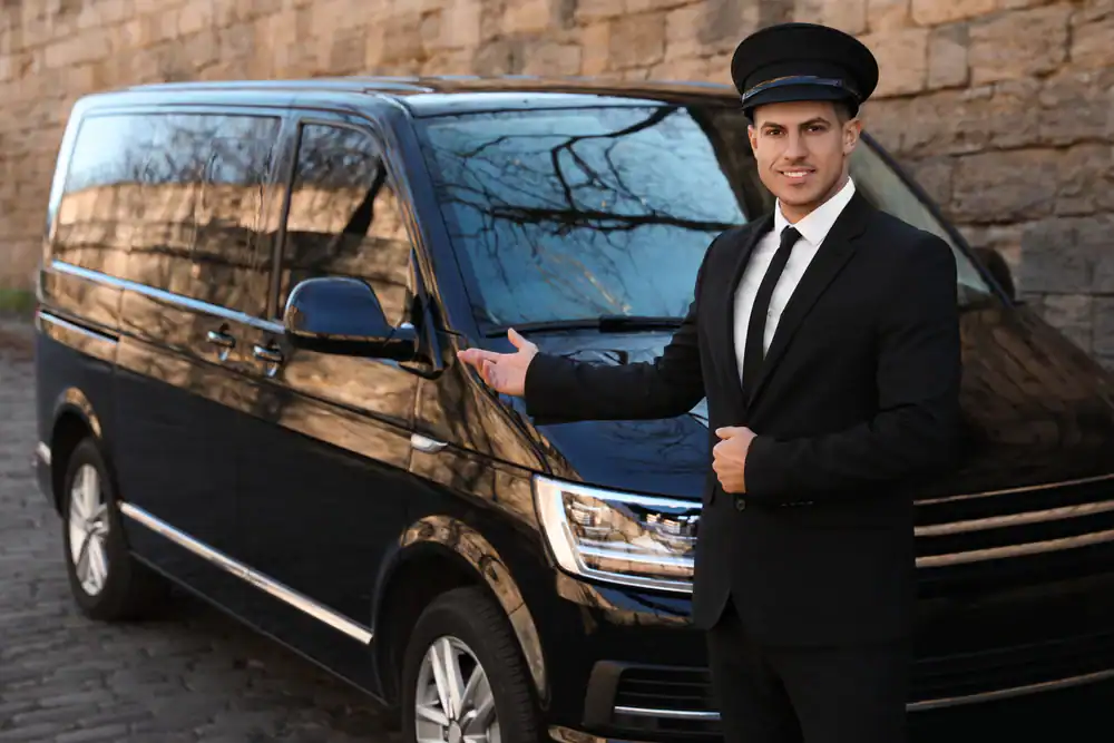 A smiling chauffeur in a black suit and cap stands next to a black van, gesturing toward the vehicle, representing premium Limo Service New Jersey, with a stone wall in the background.