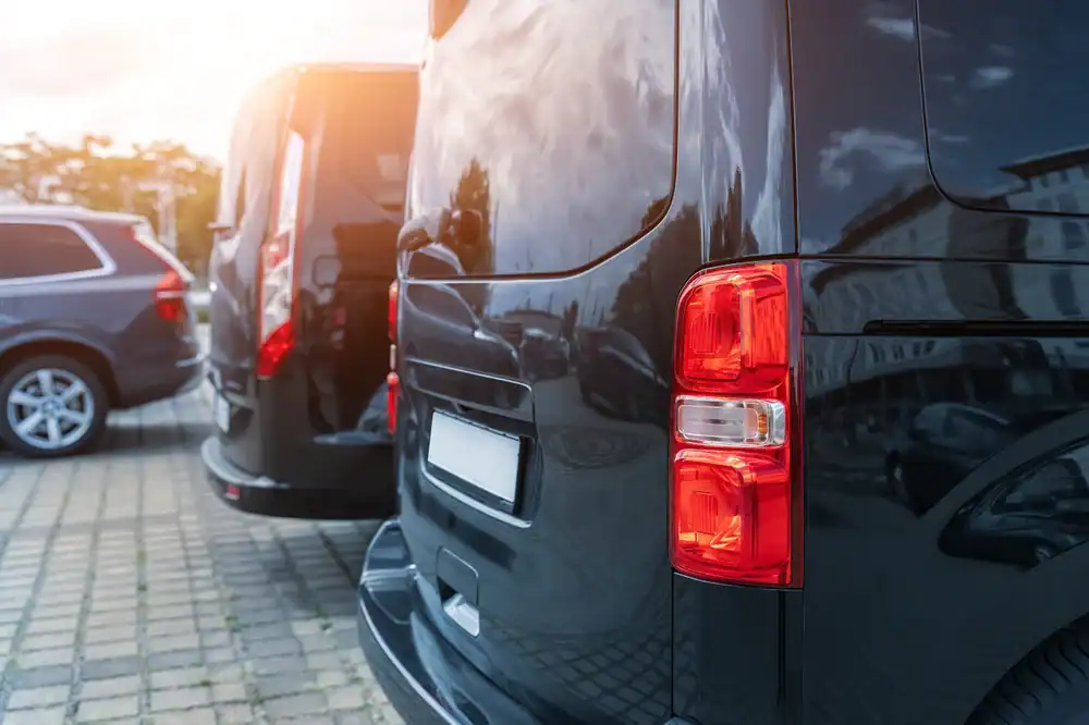 Close-up of the rear of a black van, part of a Limo Service New Jersey fleet, parked on a street with two other vehicles reflecting sunlight and nearby buildings on their shiny surfaces.