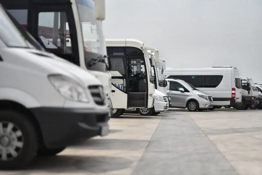 A row of white minibuses and vans, part of a Limo Service New Jersey fleet, are parked in an outdoor lot on a cloudy day, with a gray car visible among them.