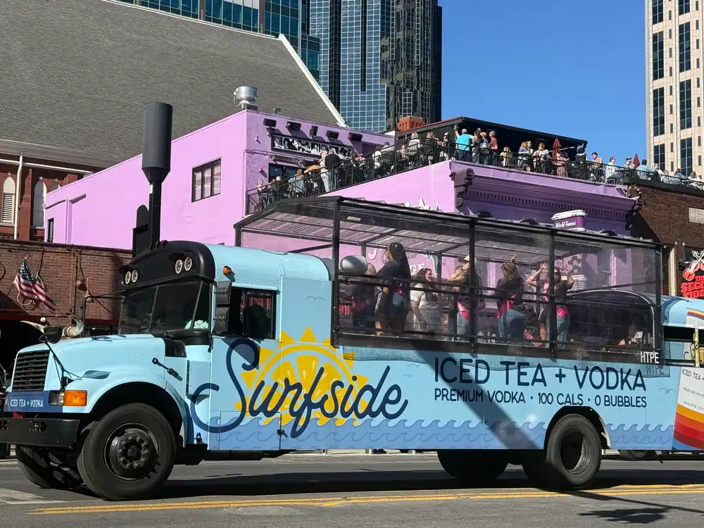 A blue bus with &ldquo;Surfside Iced Tea + Vodka&rdquo; branding, reminiscent of a Limo Service New Jersey ride, carries people dancing inside a transparent enclosure as it drives past a purple building and city skyscrapers under sunny skies.