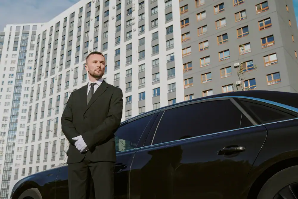A man in a black suit and white gloves stands beside a black car, representing a premium Limo Service New Jersey, in front of a tall, modern apartment building, looking to the side with a serious expression.
