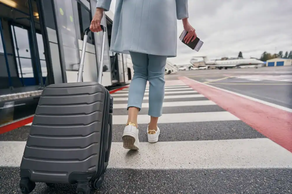 Person in light blue pants and coat walks across an airport tarmac, pulling a black suitcase and holding a passport and ticket, heading toward an airplane on a cloudy day after arriving with Limo Service New Jersey.