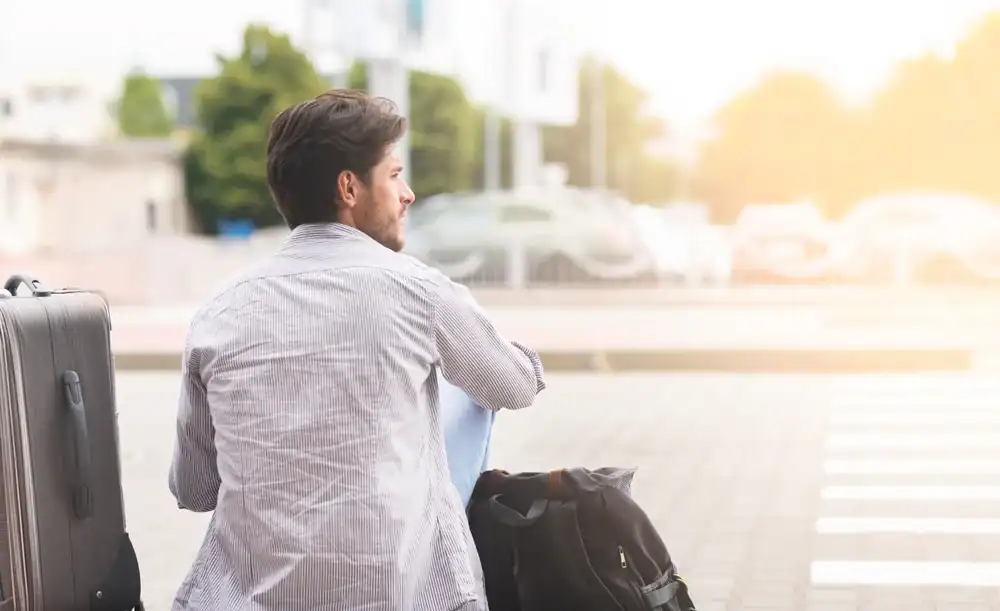 A man sits outdoors with a suitcase and a backpack, gazing into the distance. Near a street under bright sunlight, he appears to be waiting for his Limo Service New Jersey as blurred trees and buildings fill the background.