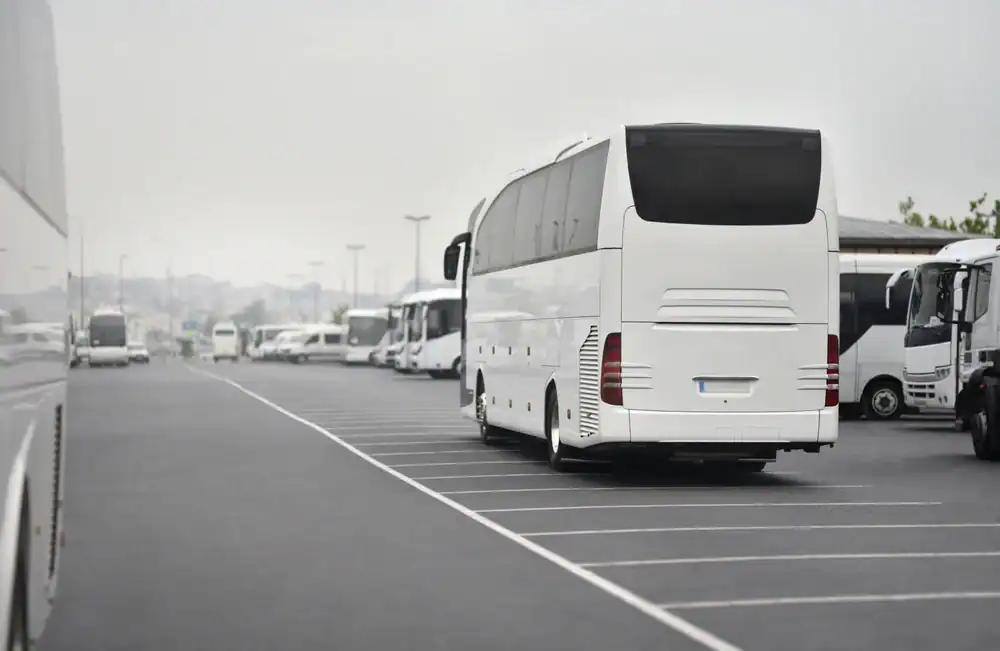 Several white buses, part of a Limo Service New Jersey fleet, are parked in an organized row in a spacious, outdoor parking lot on a cloudy day. The area appears empty except for the buses, with lines marking the parking spaces.
