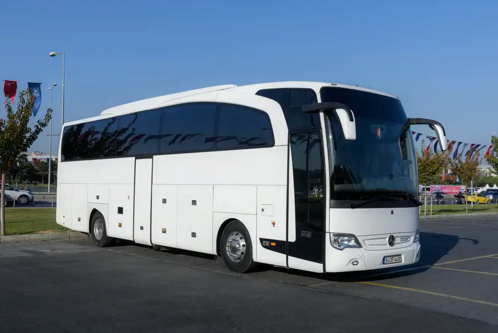 A large white coach bus, part of a premier Limo Service New Jersey, is parked in a lot near a curb, with trees and flags visible in the background on a clear day.