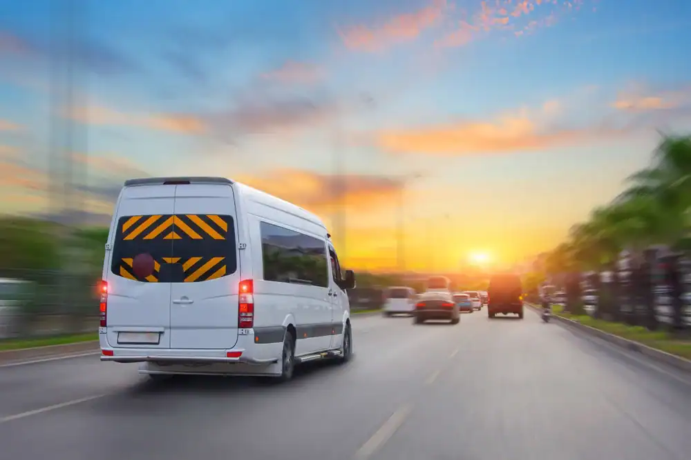 A white van driving on a city road at sunset, surrounded by other vehicles. The colorful sky with orange and blue hues adds to the sense of motion—perfect for a Limo Service New Jersey commute through the vibrant cityscape.