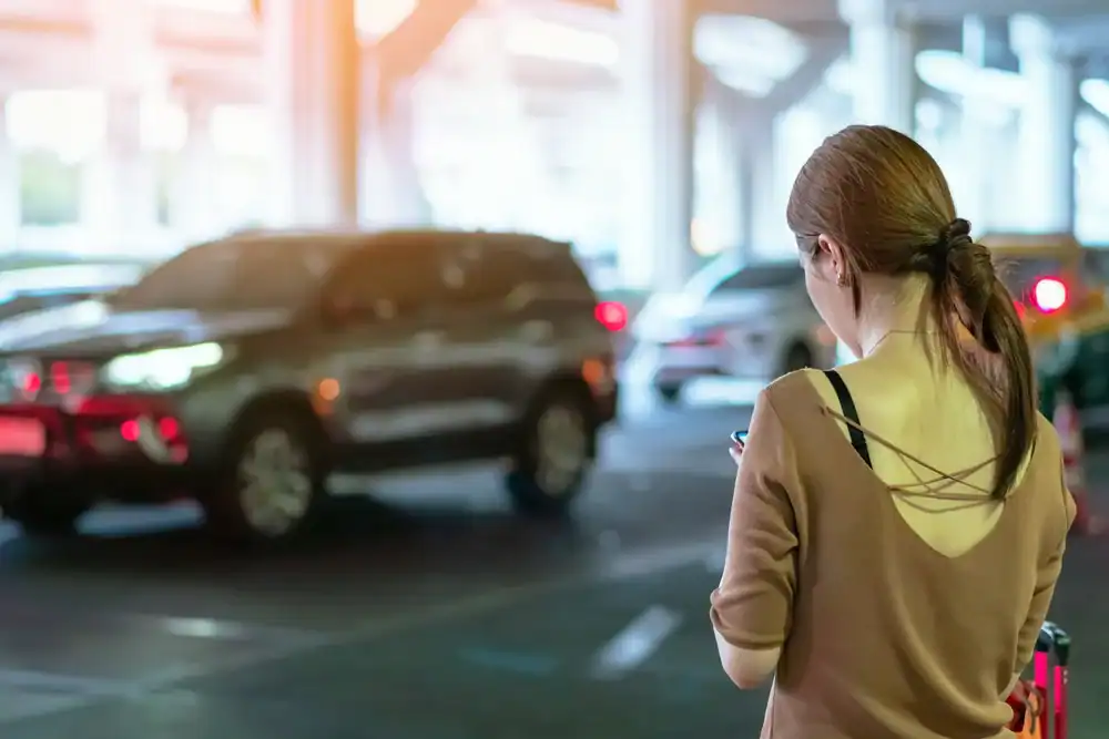 A woman with brown hair tied in a ponytail stands in a parking garage, looking at her smartphone. Cars and a Limo Service New Jersey vehicle are in the background, while sunlight filters in from the left.