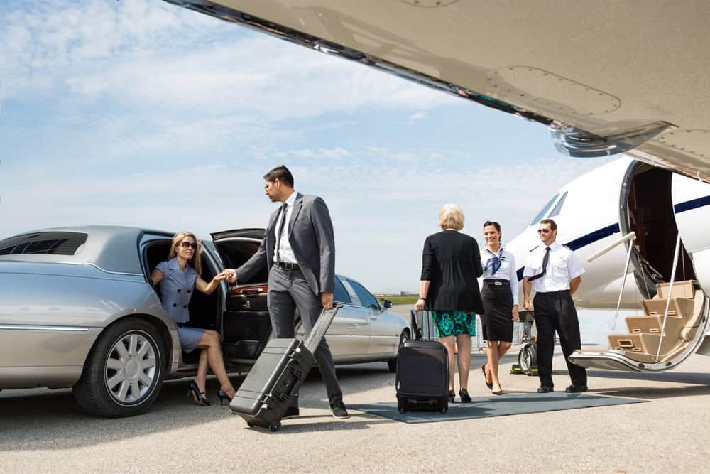 A woman in business attire exits a luxury car, provided by Limo Service New Jersey, assisted by a man with luggage. Nearby, two pilots and a flight attendant stand by a private jet as another woman approaches with her suitcase.