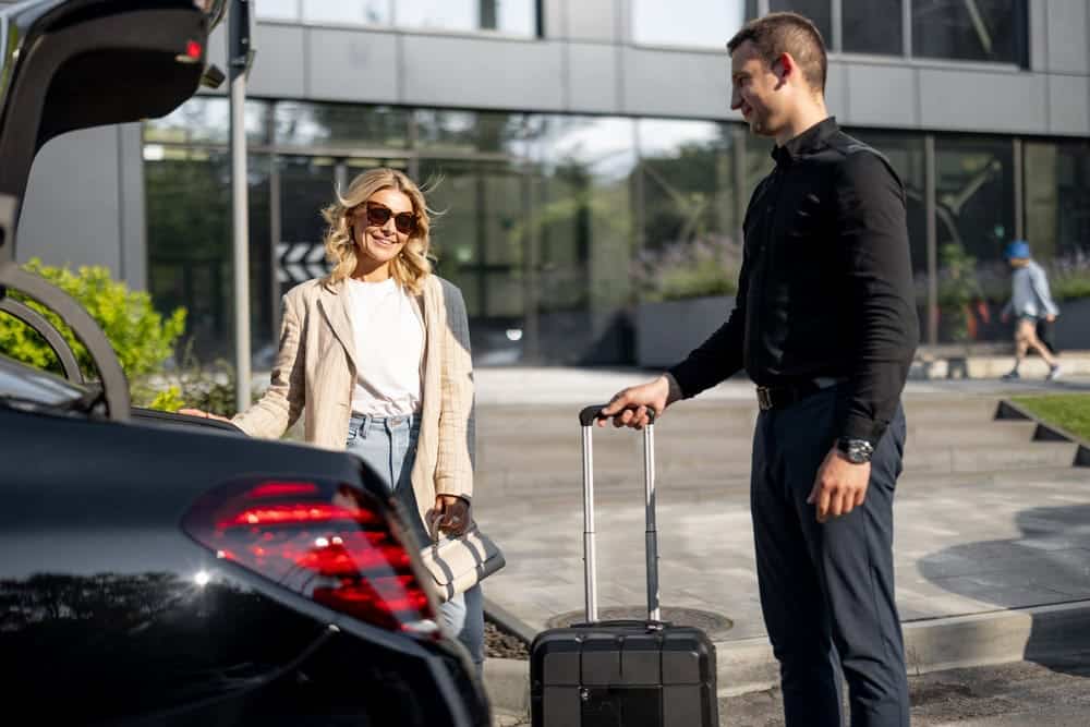 A smiling woman in sunglasses loads luggage into the trunk of a black car from a Limo Service New Jersey, while a man in business attire stands nearby holding a suitcase outside a modern building.