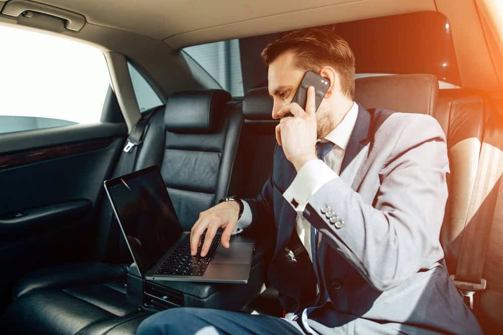 A man in a suit sits in the backseat of a Limo Service New Jersey vehicle, talking on his phone while working on a laptop placed on his lap. Sunlight streams in through the car window.